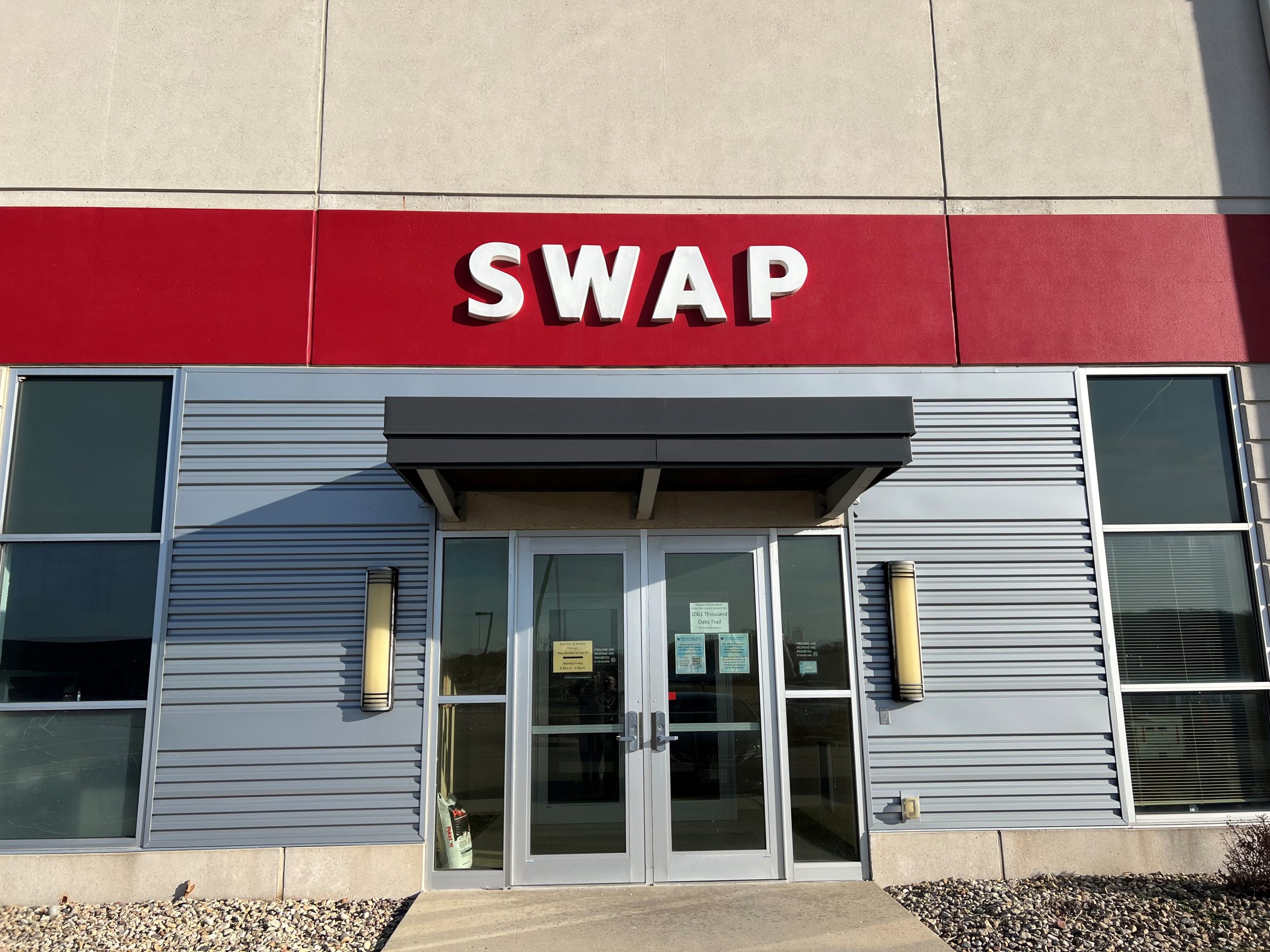 Exterior photo of the SWAP entrance. Entrance doors to a semi-modern tan cement and silver metal building façade. A dark red painted area above the doors features large white text reading "SWAP."